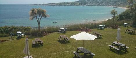 A grassy outdoor area with picnic tables and umbrellas, overlooking a coastal view with boats and a beach.