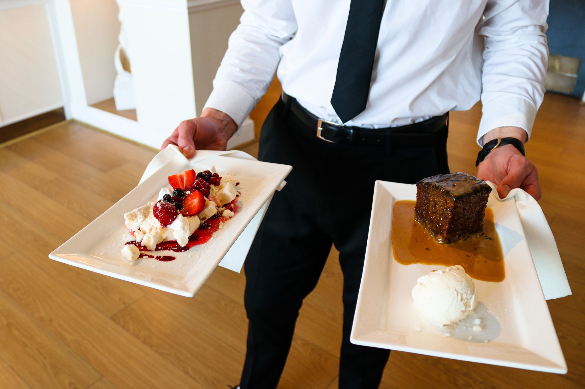 A person holding two dessert plates, one with a fruit-topped dessert and the other with a dessert and ice cream.