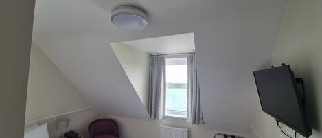 Habitación de hotel con cama, una silla, un escritorio, televisor, ventana con cortinas, radiador y una lámpara en el techo.