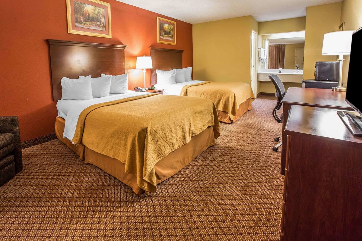 A hotel room with two beds, a desk, a chair, a TV, and a bathroom visible through an open door.