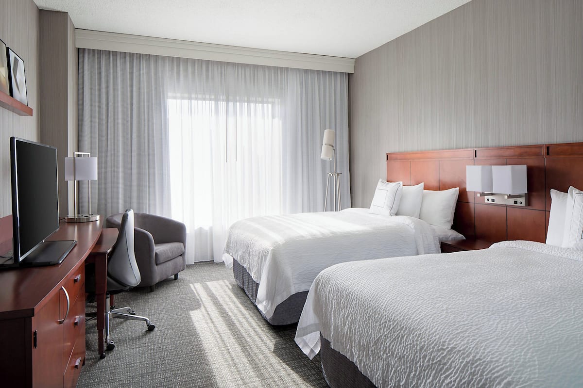 A hotel room with two beds, a desk with a TV, a chair, and a large window with curtains.