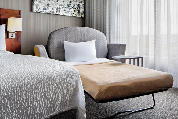 Una habitación de hotel con una cama, una mesita de noche con lámpara, una silla y vistas a una ventana con cortinas.