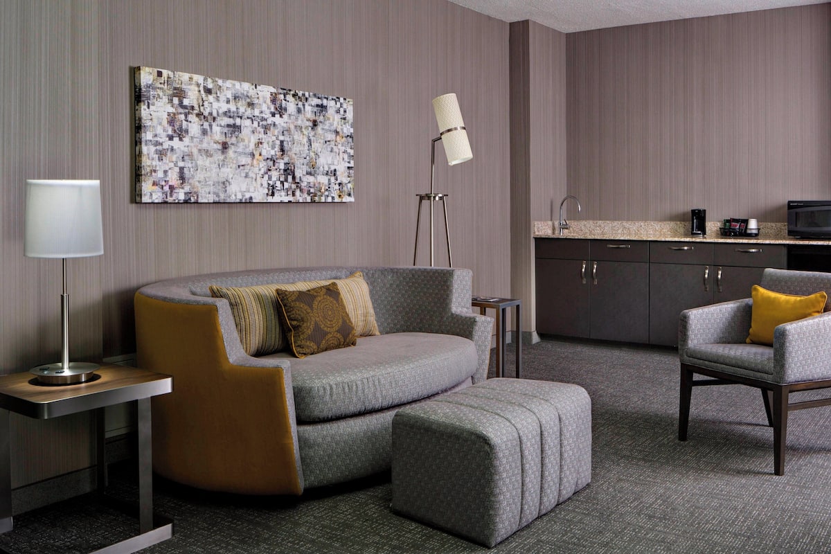 A modern hotel room with a grey sofa, ottoman, and armchair, a small side table, a floor lamp, a kitchen area with a microwave, and a large abstract painting on the wall.