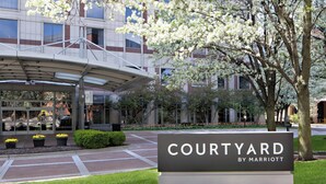 Señal de Courtyard by Marriott con un acceso cubierto, una zona ajardinada con árboles y arbustos, y un edificio de estilo arquitectónico moderno al fondo.