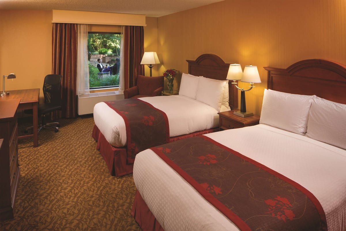 Quarto de hotel com duas camas, uma escrivaninha e uma cadeira. Há uma janela com cortinas, um abajur e um vaso com flores.
