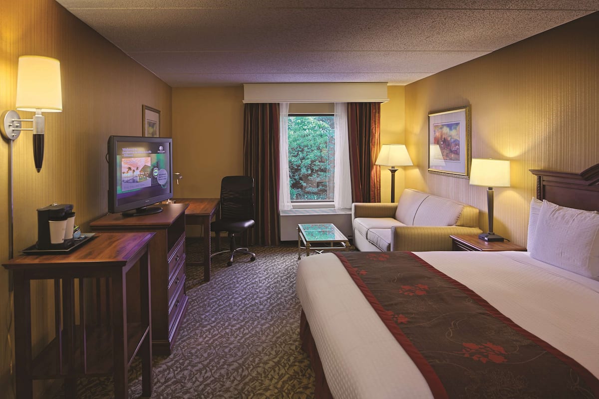 Quarto de hotel com cama, escrivaninha, cadeira, televisão e luminárias.
