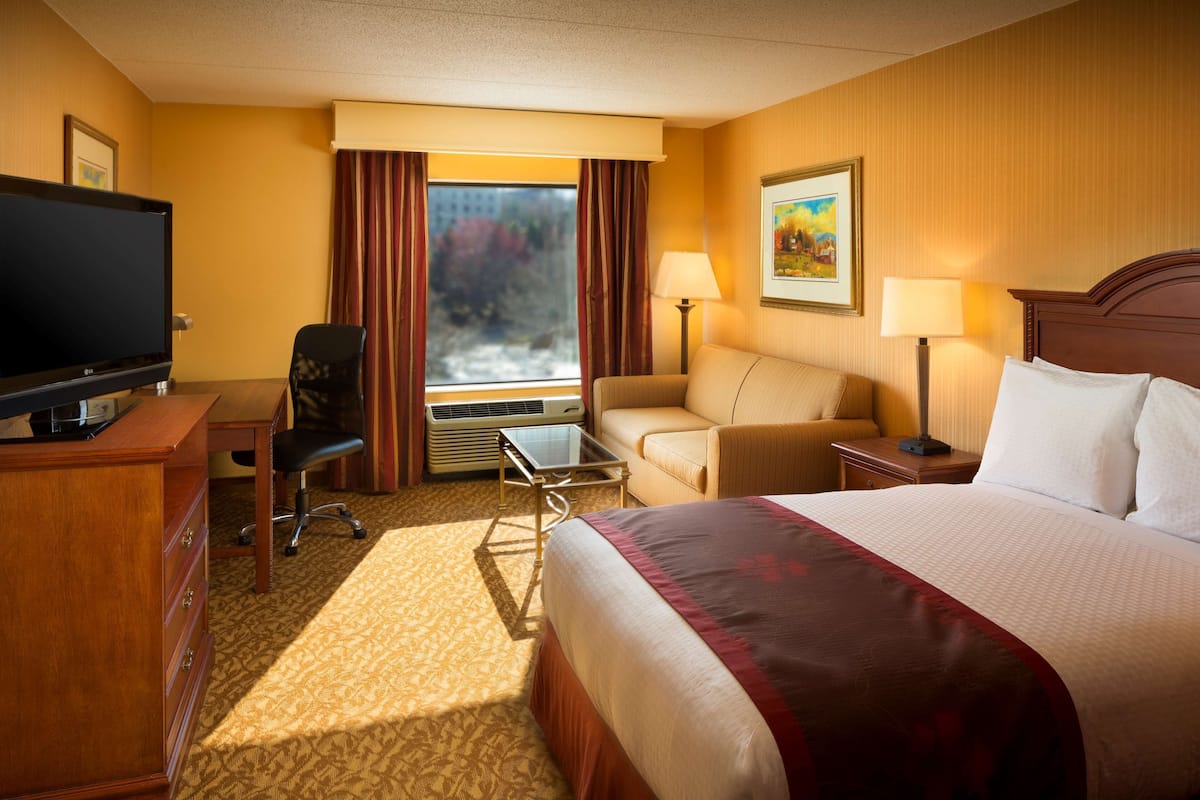 Quarto de hotel com cama, secretária com computador, cadeira, televisão, janela com cortinas e um quadro na parede.