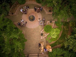 An aerial view of a circular outdoor seating area with tables and chairs, surrounded by landscaped gardens and trees.