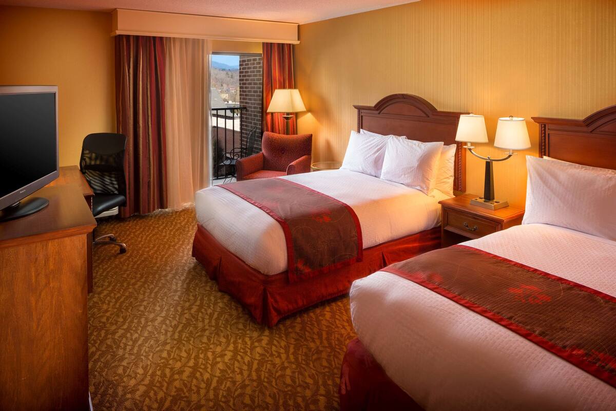 Quarto de hotel com duas camas, televisão, escrivaninha e varanda com cortinas.