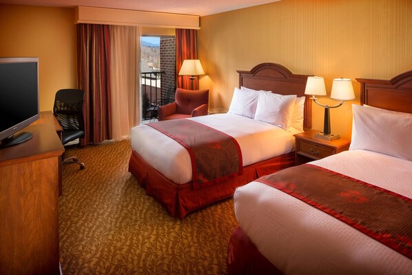 A hotel room with two beds, a TV, a desk, and a balcony with curtains.