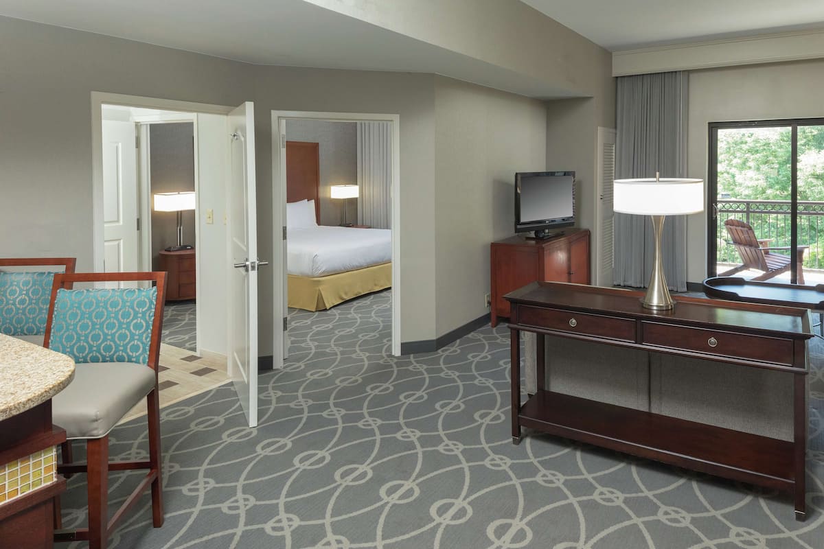Quarto de hotel com cama, escrivaninha com luminária, televisão, cadeira e vista para o exterior.