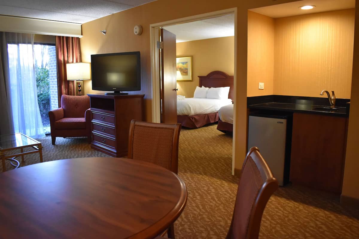 Quarto de hotel com mesa de jantar, cadeiras, televisão, cama, pequeno espaço de cozinha e banheiro.