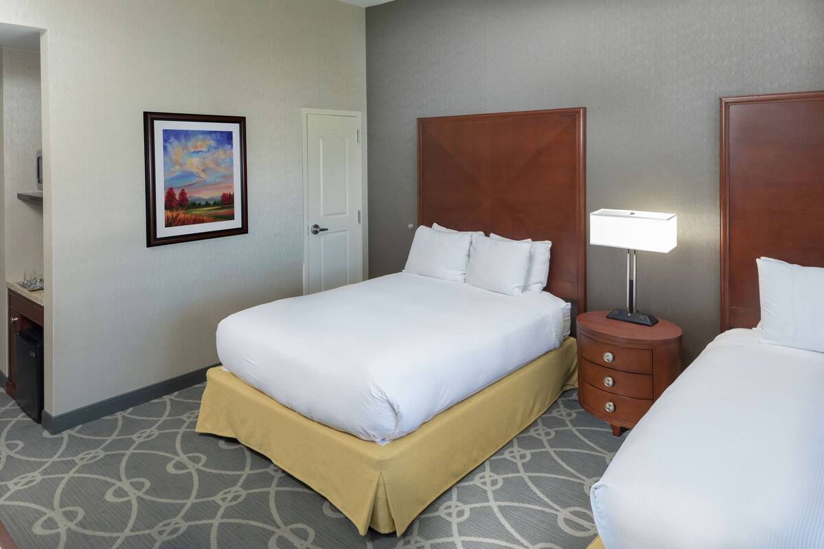 Quarto de hotel com uma cama grande, uma mesa de cabeceira com abajur, um quadro na parede e uma porta.