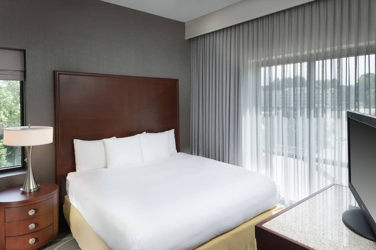 Quarto de hotel com uma cama grande, mesa de cabeceira com abajur, televisão e janela com cortinas.