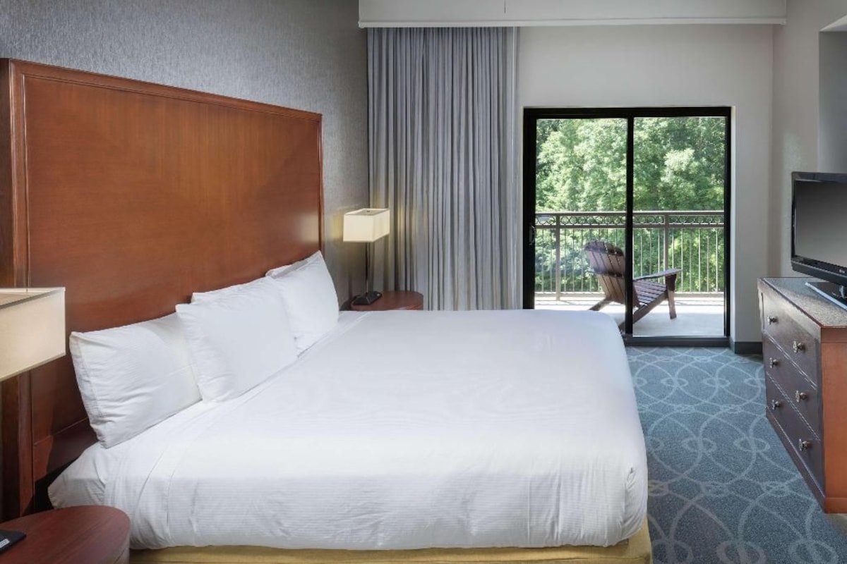 A hotel room with a large bed, a TV, a desk, and a balcony with a chair.