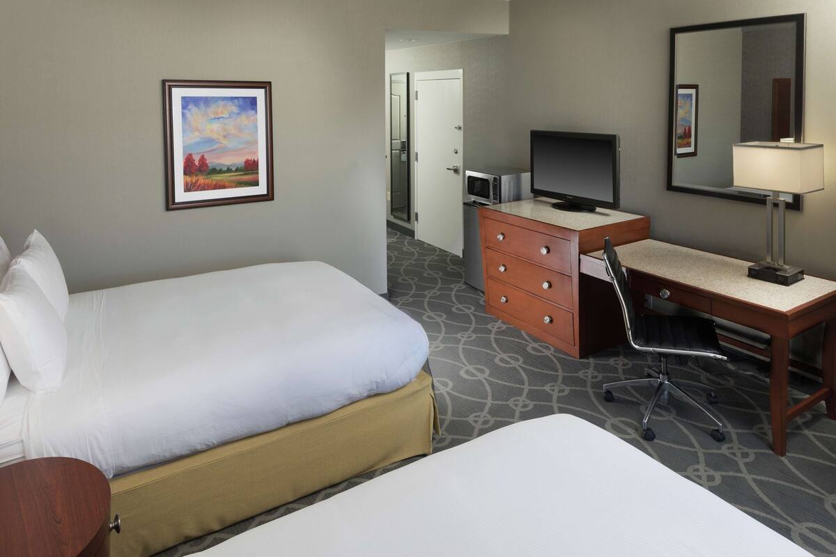 Quarto de hotel com duas camas, uma escrivaninha com computador, televisão, espelho e um quadro na parede.
