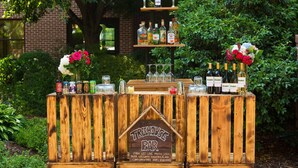 Eine Holztresore mit einer Beschilderung mit dem Aufschrift "Treehouse Bar", präsentiert eine Auswahl an alkoholischen Getränken, Gläsern und dekorativen Blumen.