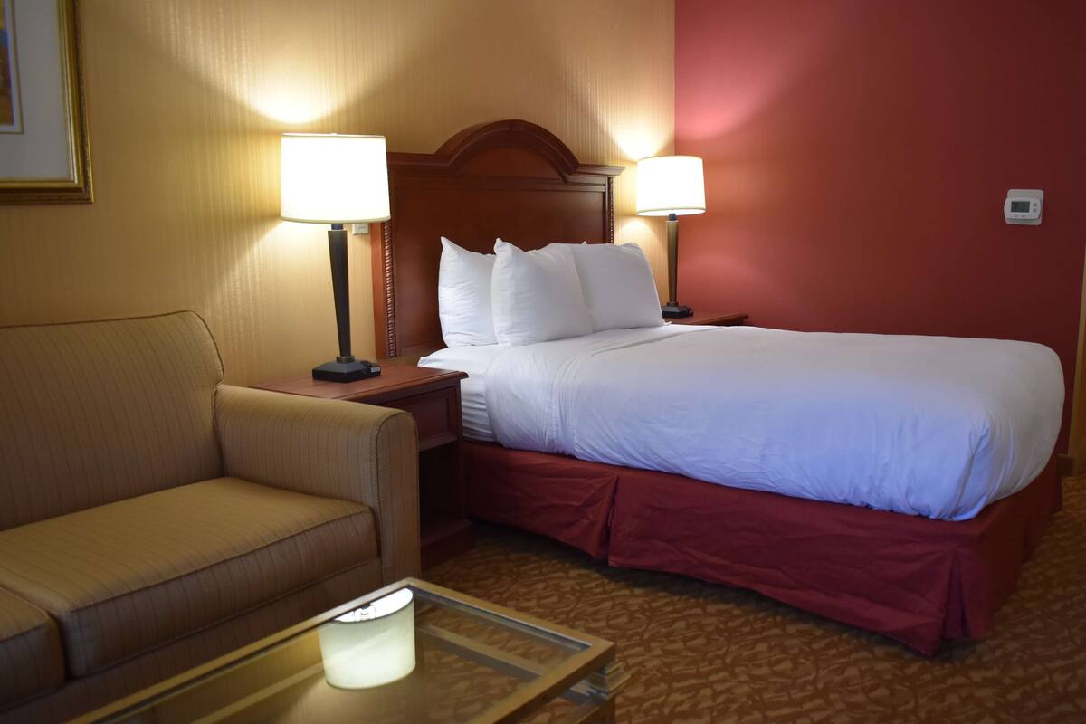Quarto de hotel com cama, sofá, duas luminárias de cabeceira e mesa de apoio.