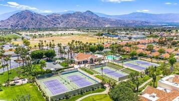 A tennis court complex with multiple courts, surrounded by residential buildings and palm trees, with mountains in the background.