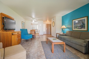 A living room with a television, a blue armchair, a sofa, a coffee table, and a painting on the wall. - Desert Breezes Resort (Palm Desert)