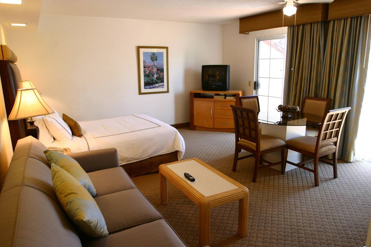 A hotel room with a bed, a sofa, a small table, a TV, a desk with chairs, and a window with curtains.
