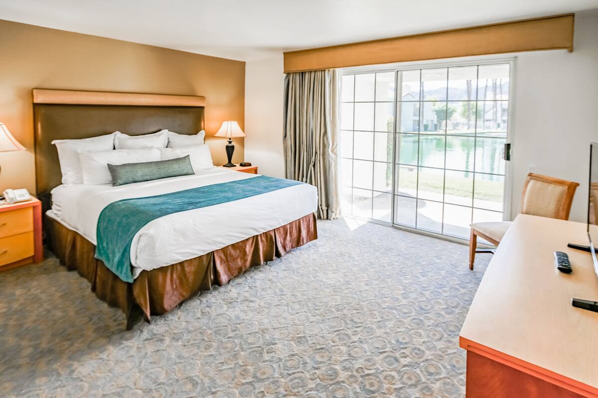 A hotel room with a large bed, a desk, a chair, a telephone, and a window with a view of a pool and greenery.