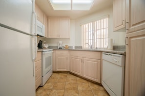 A compact kitchen with white appliances, a window with vertical blinds, and a tiled floor. - Desert Breezes Resort (Palm Desert)