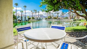Un patio con una mesa redonda y sillas a rayas azules, con vistas a una piscina con una fuente, rodeado de palmeras y edificios.