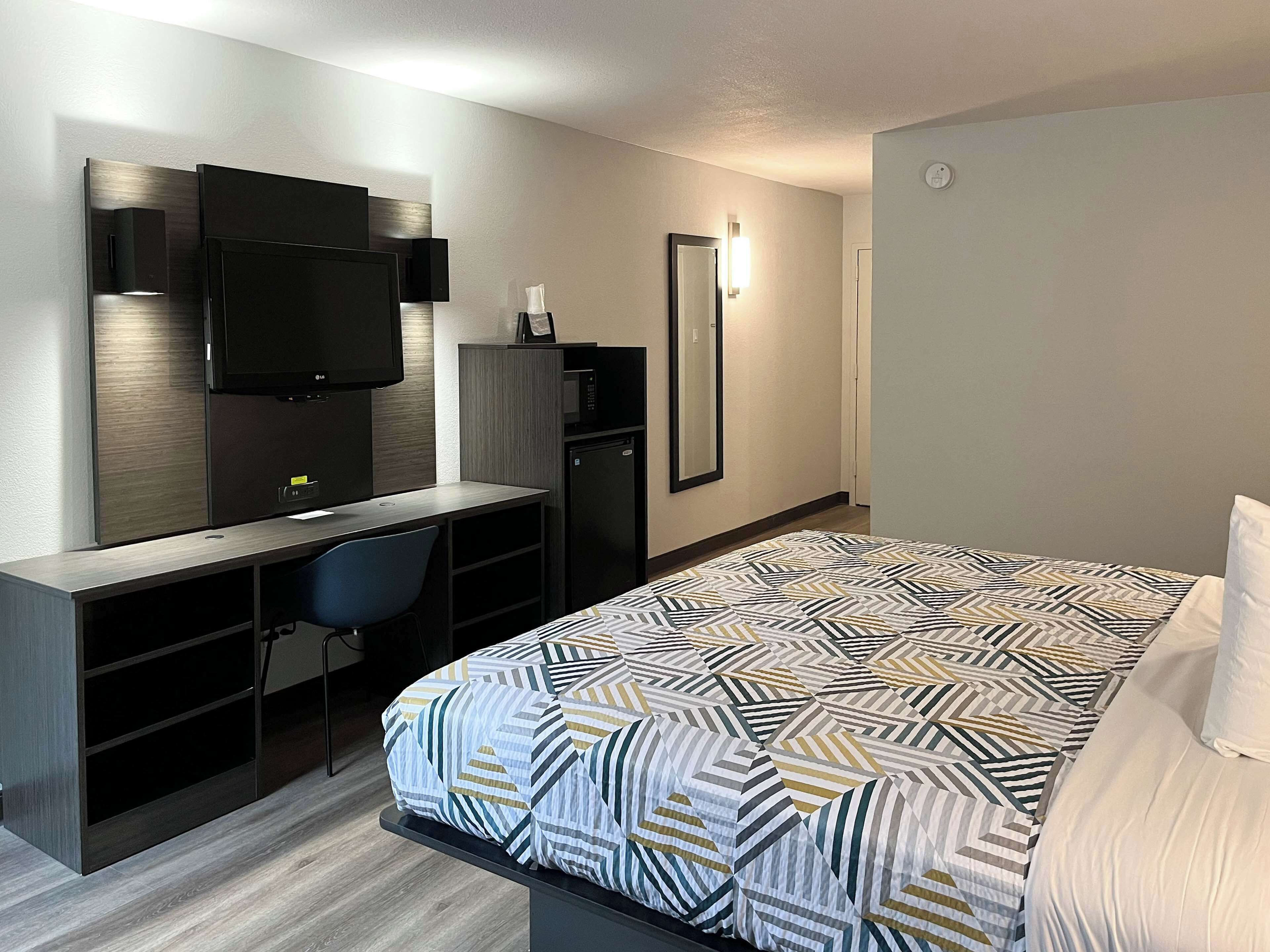 Una habitación de hotel moderna con una cama, un escritorio con una silla, un televisor y un espejo.