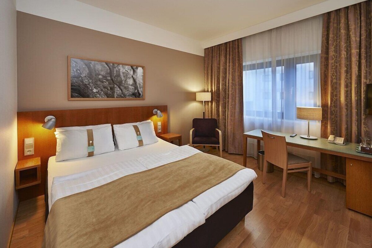 A hotel room with a large bed, a desk, a chair, and a window with curtains.