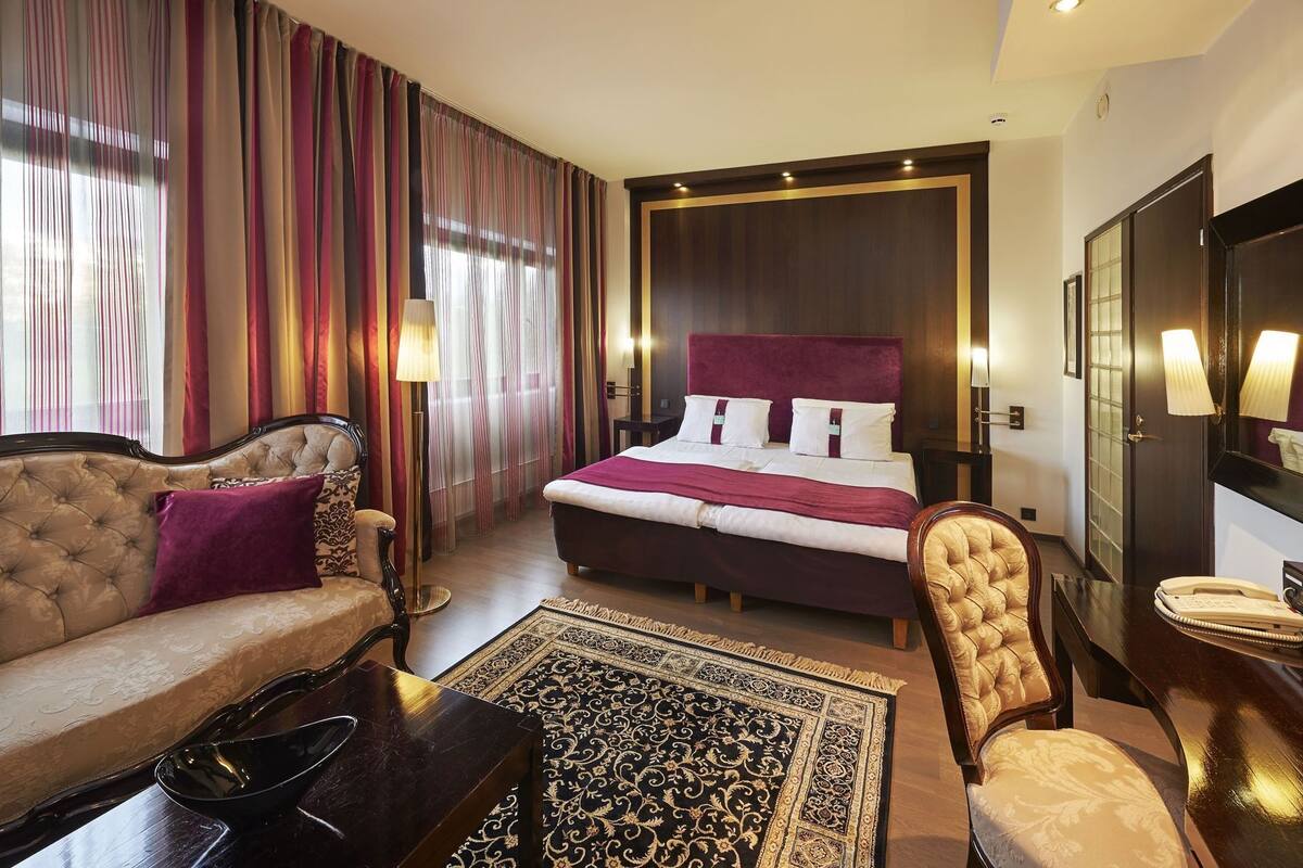 A hotel room with a large bed, a sofa, a chair, a desk, and a nightstand.