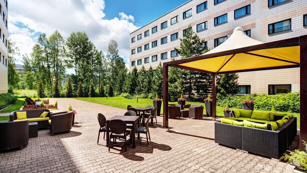 An outdoor patio with wicker furniture and a yellow canopy, surrounded by a landscaped garden and a hotel building. - Scandic Veromies (Vantaa)