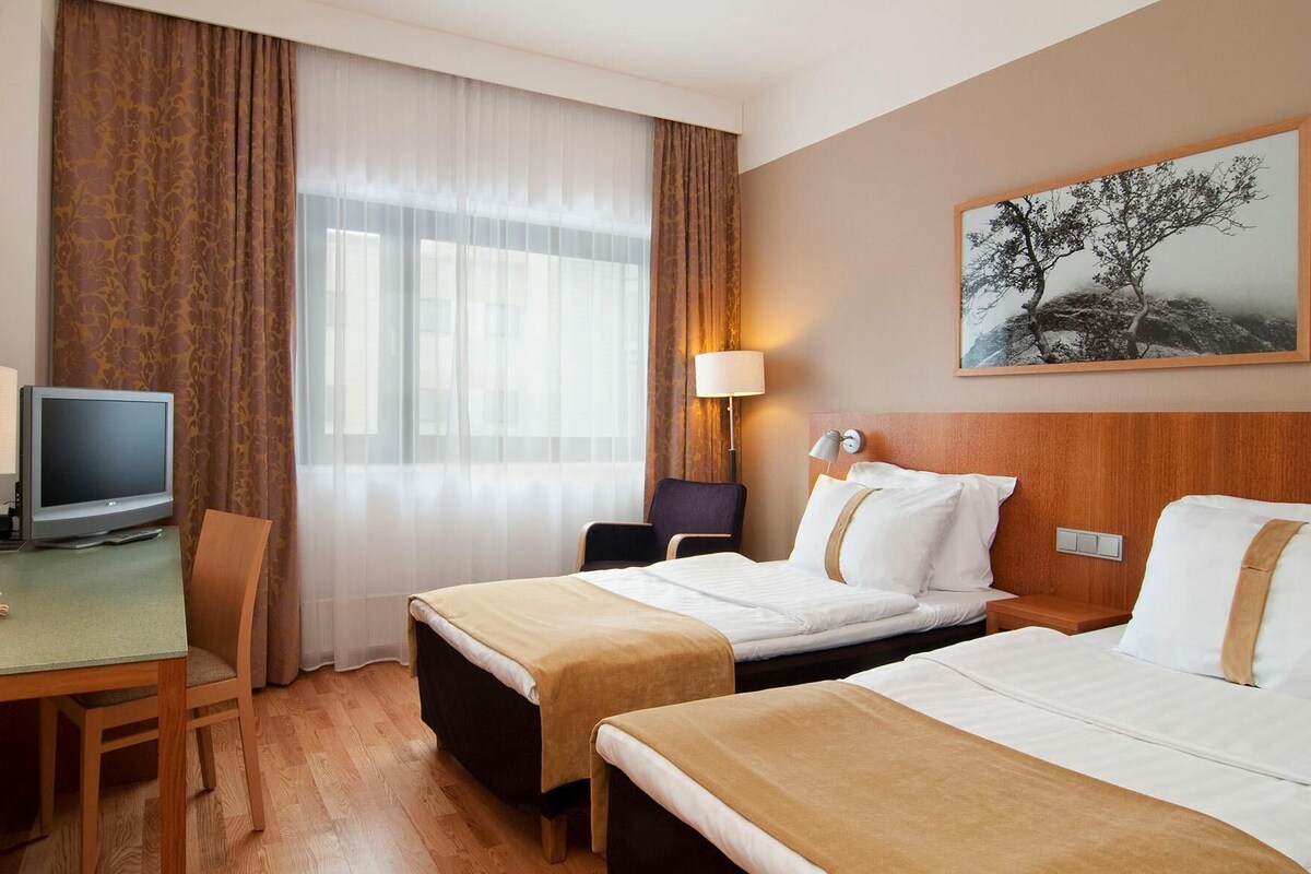 A hotel room with two beds, a desk with a television, a chair, a lamp, and a painting on the wall.