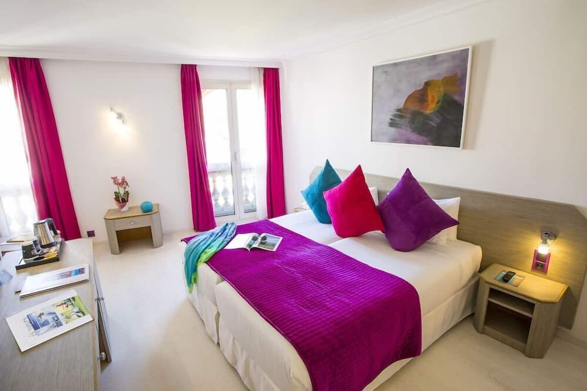 A hotel room with a bed, a nightstand, a desk, a window with curtains, and a painting on the wall.