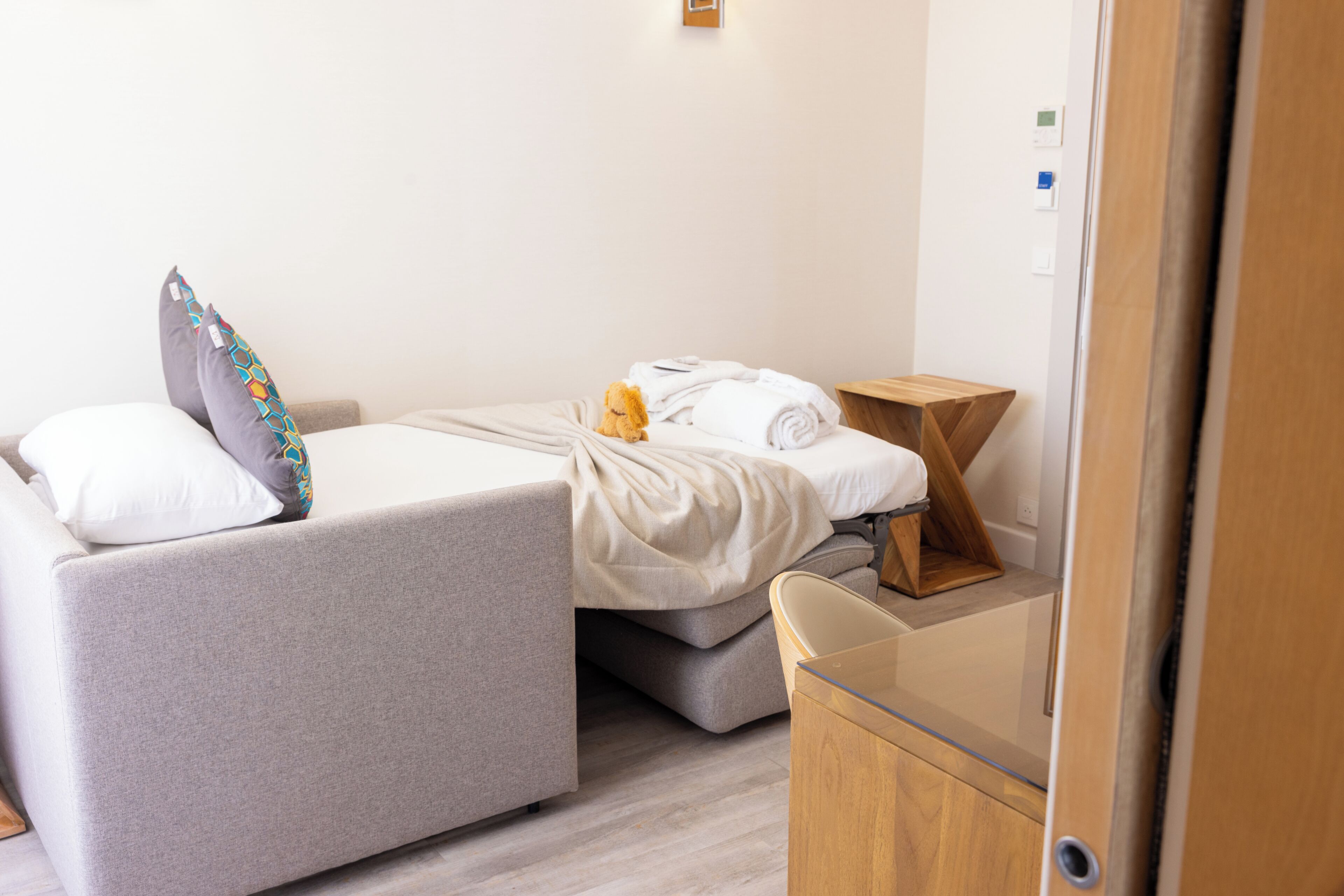 Quarto de hotel moderno com uma cama de solteiro, uma pequena mesa de cabeceira e uma mesinha de madeira.