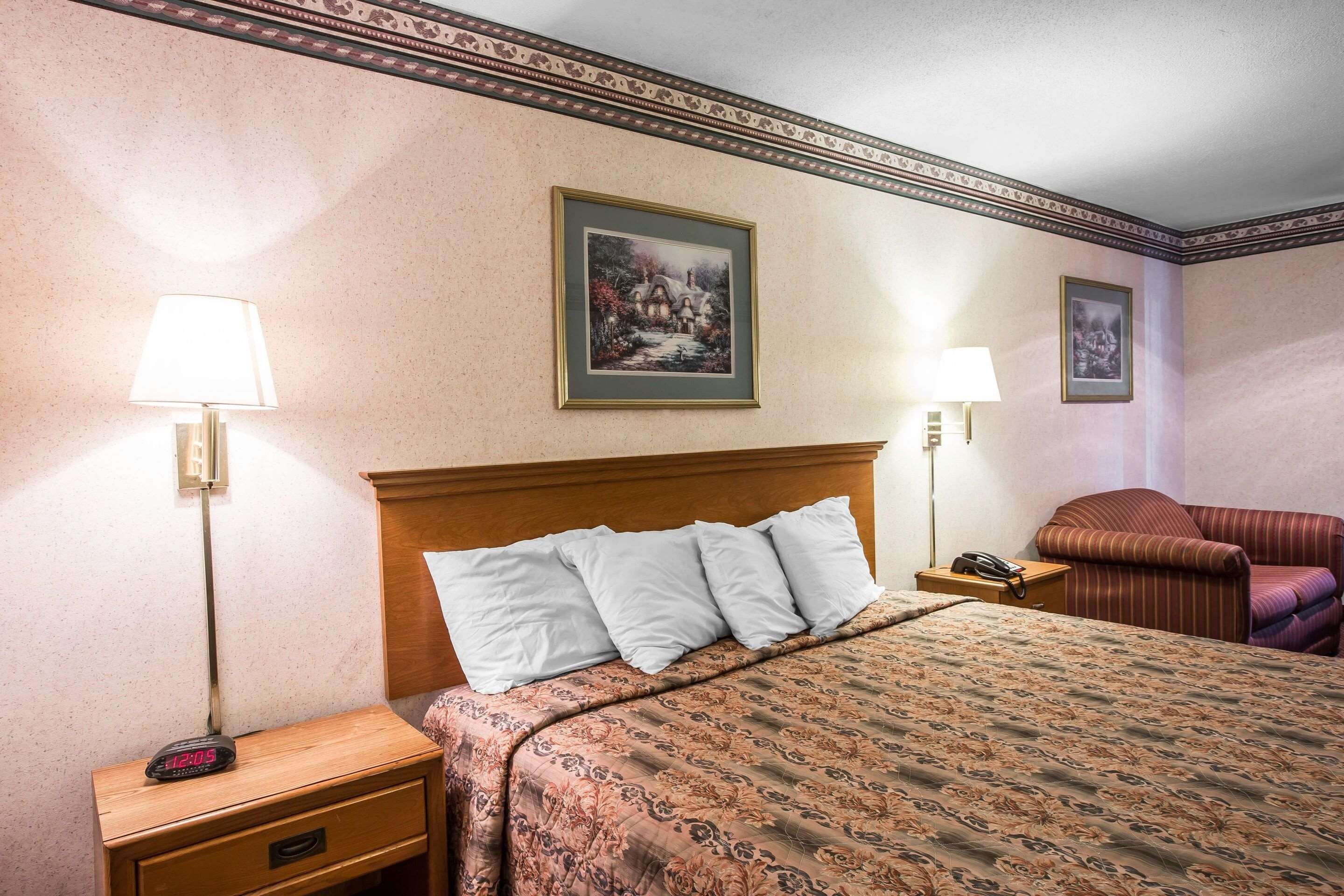 A hotel room with a bed, two bedside lamps, a nightstand with a clock, a telephone, and a chair.