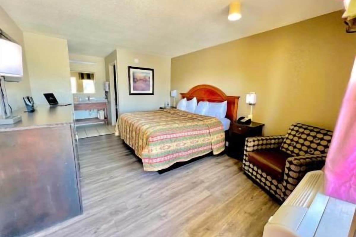 Quarto de hotel com cama, mesa de cabeceira, luminária e vista para o corredor.