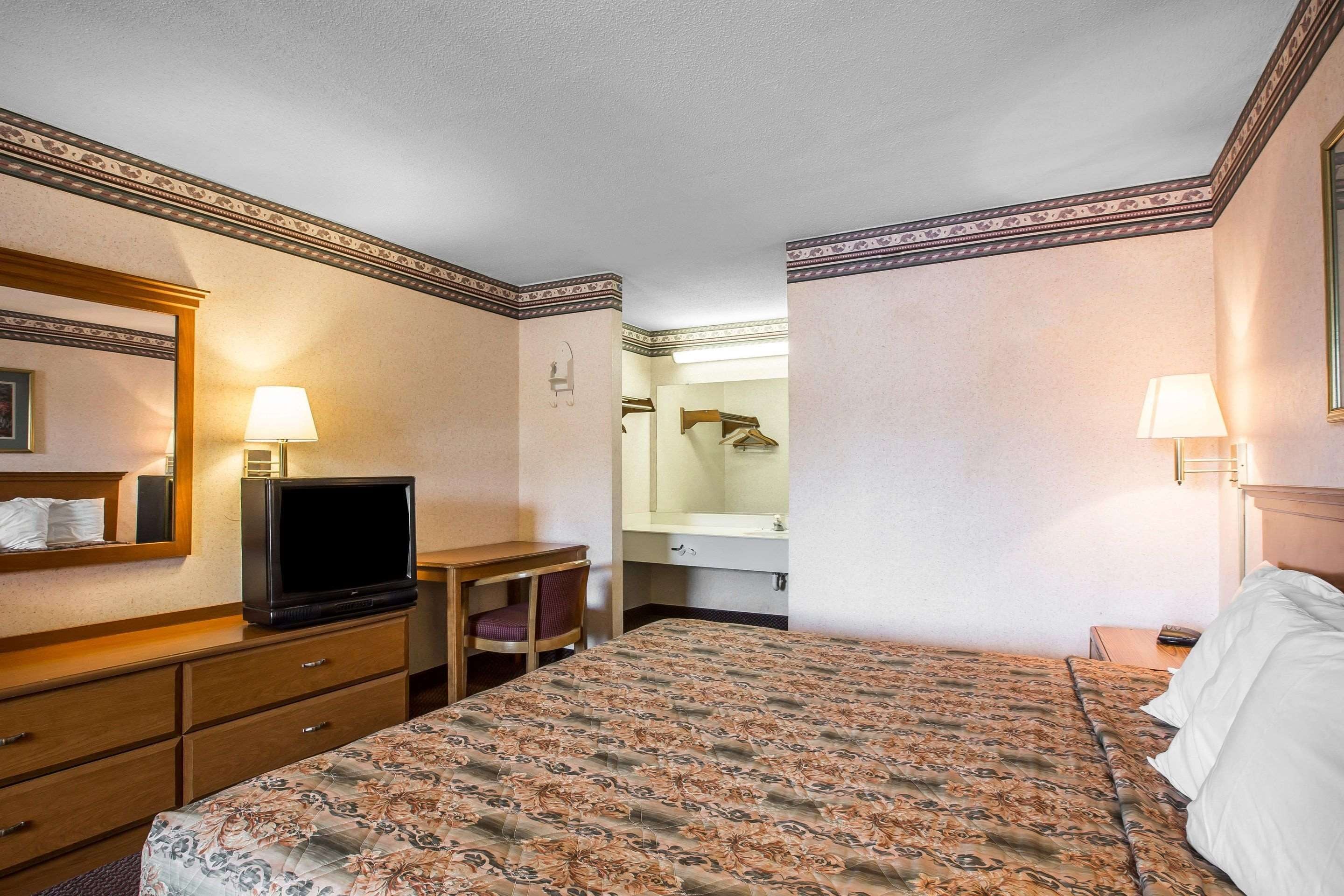 A hotel room with a bed, a desk with a television, a mirror, and a bathroom visible through an open door.