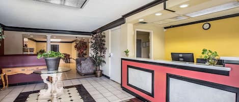 Uma área de recepção de hotel com balcão de check-in, um espaço de estar com mesa e cadeiras, e um corredor que dá acesso a outros quartos.