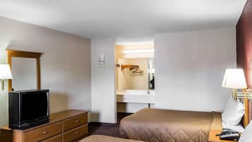 Ein Hotelzimmer mit Fernseher auf einer Holzkombie, einem Bett mit gestepptem Kopfteil, einem Nachttisch mit Lampe und einem durchgegangene Tür sichtbaren Badezimmer.