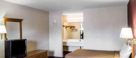 Une chambre d’hôtel avec un téléviseur posé sur une commode en bois, un lit doté d’une tête de lit matelassée, une table de chevet avec une lampe, et une salle de bain visible à travers une porte ouverte.
