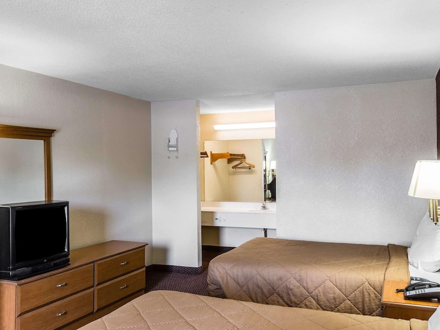 Een hotelkamer met een televisie op een houten dressoir, een bed met een gewatteerd hoofdbord, een nachtkastje met een lamp en een badkamer zichtbaar door een geopende deur.