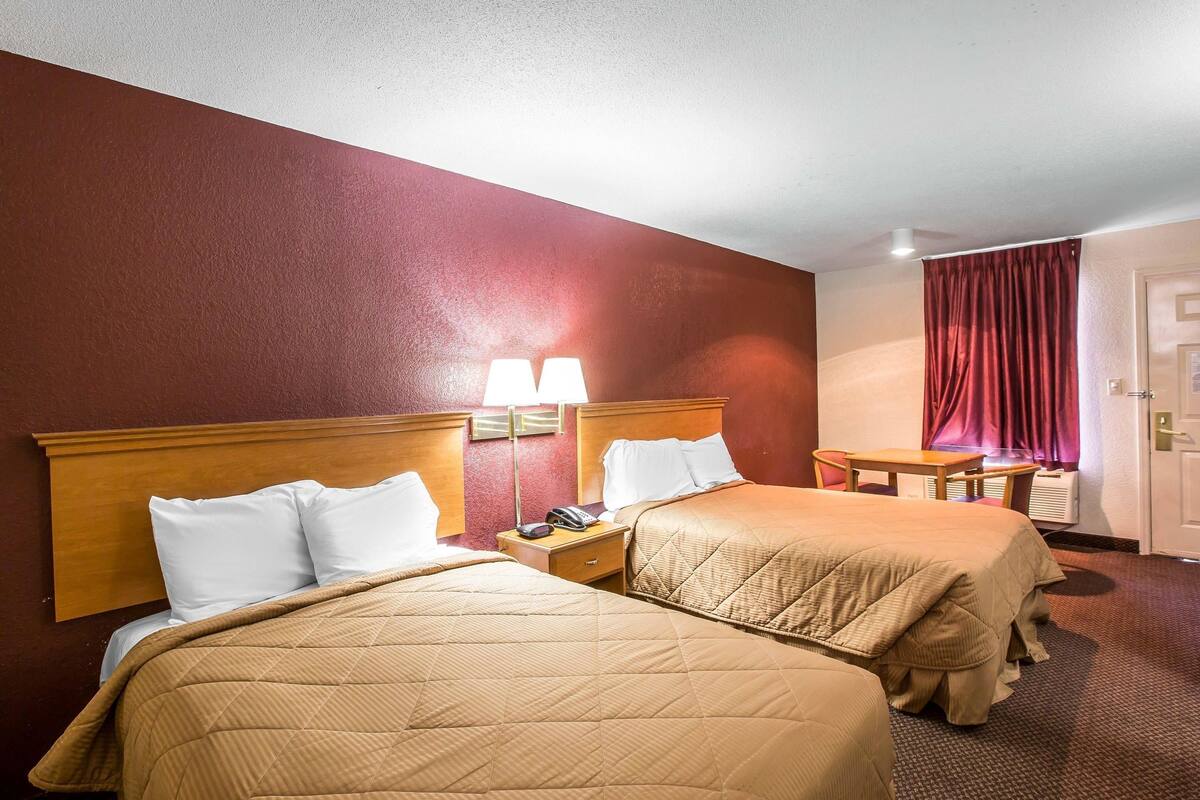 A hotel room with two beds, a nightstand with a lamp, a table, and a window with curtains.