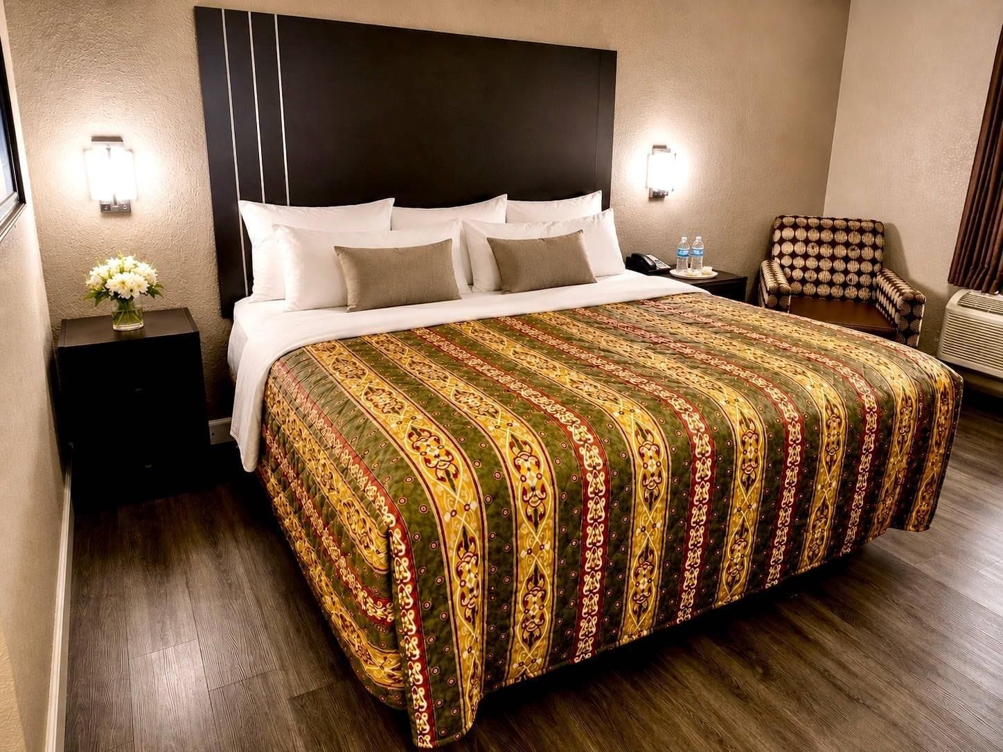 A hotel room with a large bed, a nightstand with a vase of flowers, a chair, and a wall-mounted air conditioner.