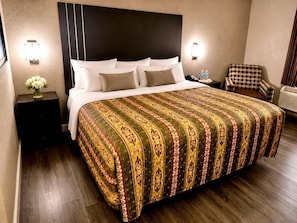 A hotel room with a large bed, a nightstand with a vase of flowers, a chair, and a wall-mounted air conditioner. - Rodeway Inn Expo Center (Spartanburg)