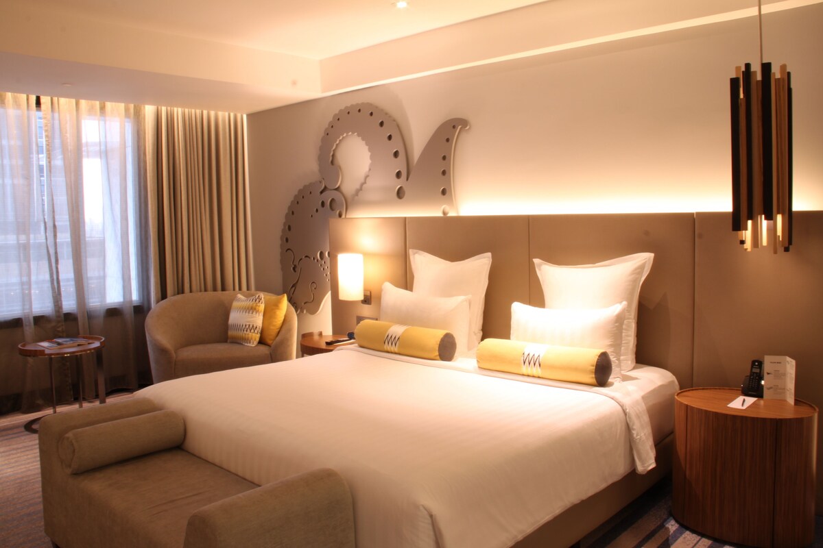 Habitación de hotel con una cama grande, un sofá, una mesita de noche y una lámpara.