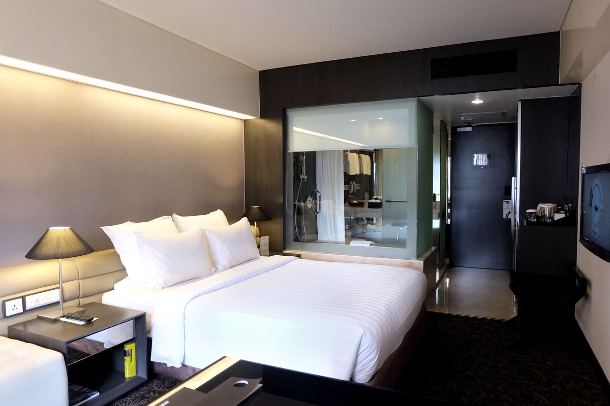 Habitación de hotel moderna con una cama grande, una mesita de noche con lámpara, un espejo y un televisor.