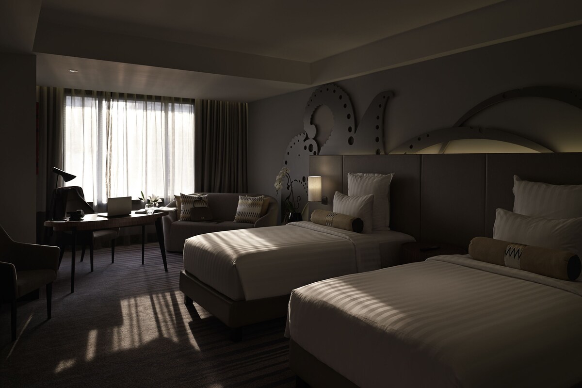 Habitación de hotel con dos camas, un escritorio, una silla y un ventanal con cortinas.