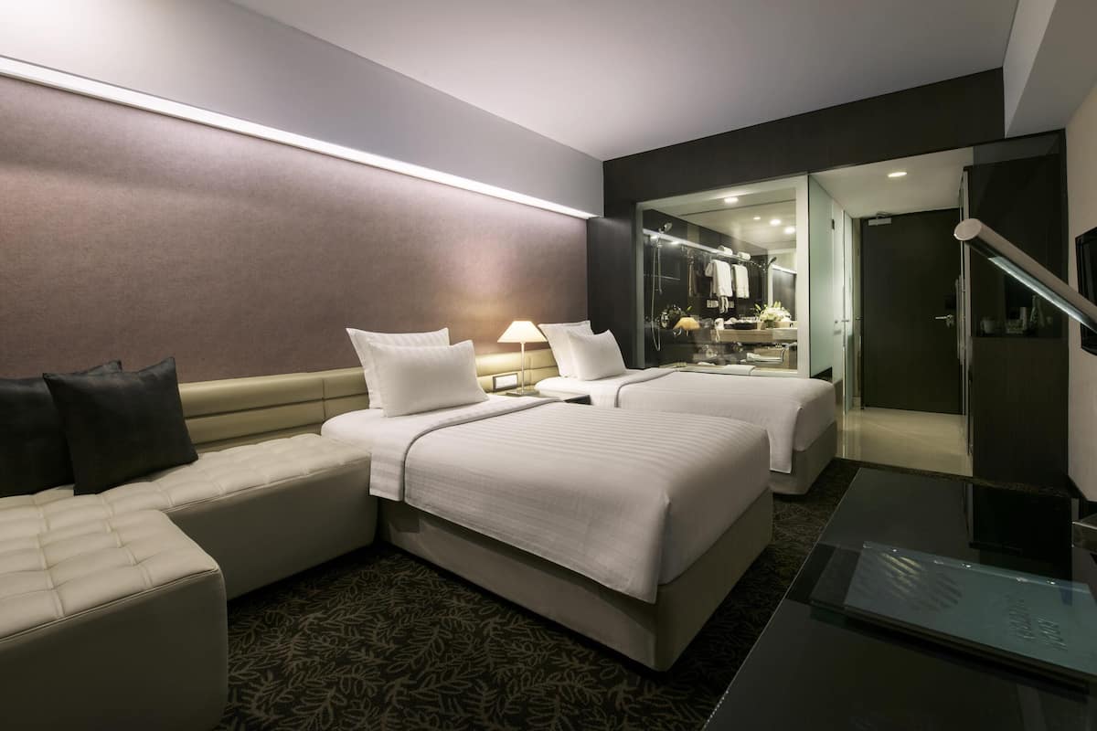 A hotel room with two beds, a sofa, a glass shower enclosure, and a separate bathroom visible through a glass door.
