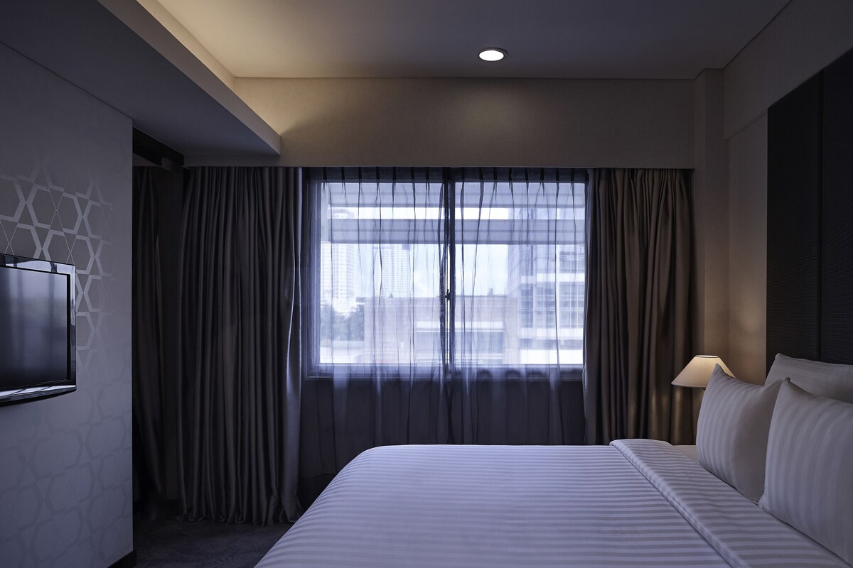 Una habitación de hotel con una cama, un televisor montado en la pared, una ventana con cortinas y una lámpara en la mesita de noche.